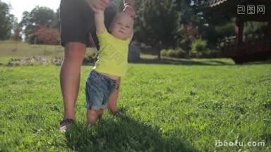 可爱的蓝眼睛蹒跚学步的男孩在父亲的帮助下在夏季公园的绿色草地上迈出第一步的肖像